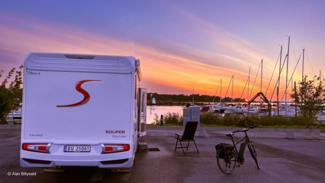 Dette er et bilde som viser en hvit bobil parkert ved siden av en rolig vannkant, sannsynligvis en marina eller en innsjø, ved solnedgang.