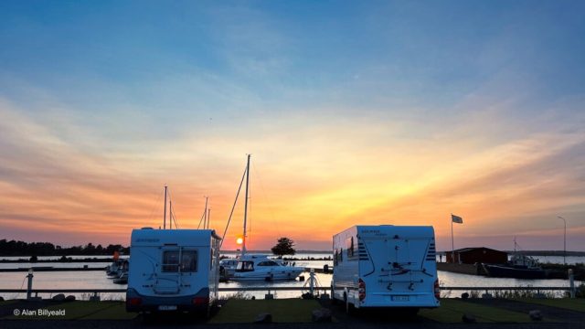 Bildet viser to hvite bobiler parkert ved siden av hverandre på en asfaltert parkeringsplass ved en marina. Det er tatt et bilde under en vakker solnedgang, som gir himmelen en fargerik blanding av oransje, rosa og blå toner.