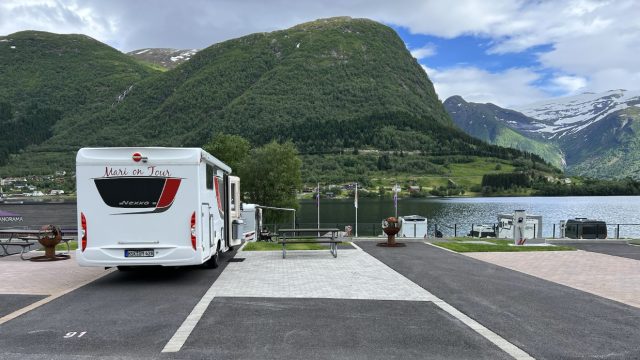 Bidet viser en hvit bobil parkert på en asfaltert parkeringsplass med en spektakulær utsikt over et vann og fjellandskap.
