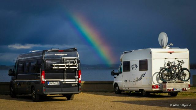 To bobiler som er parkert ved siden av hverandre på en parkeringsplass. Det er et unikt øyeblikksbilde, da det er et regnbue som strekker seg over himmelen i bakgrunnen.